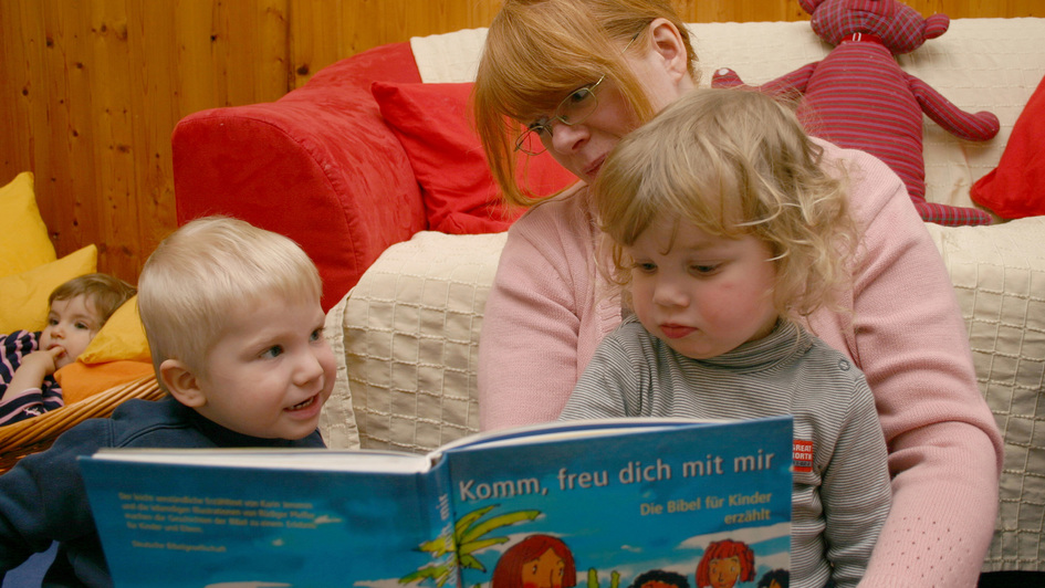 Eine Erzieherin hat zwei beiden Zweijährige auf dem Schoß und liest ihnen aus einer Kinderbeibel vor
