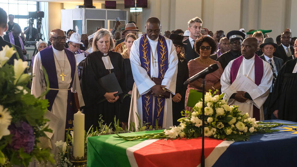 Vertreter der Kirchen in Namibia, der EKD, der Republik Namibia und der Bundesregierung stehen an einem Sarg. der mit der namibischen Fahne bedeckt ist.