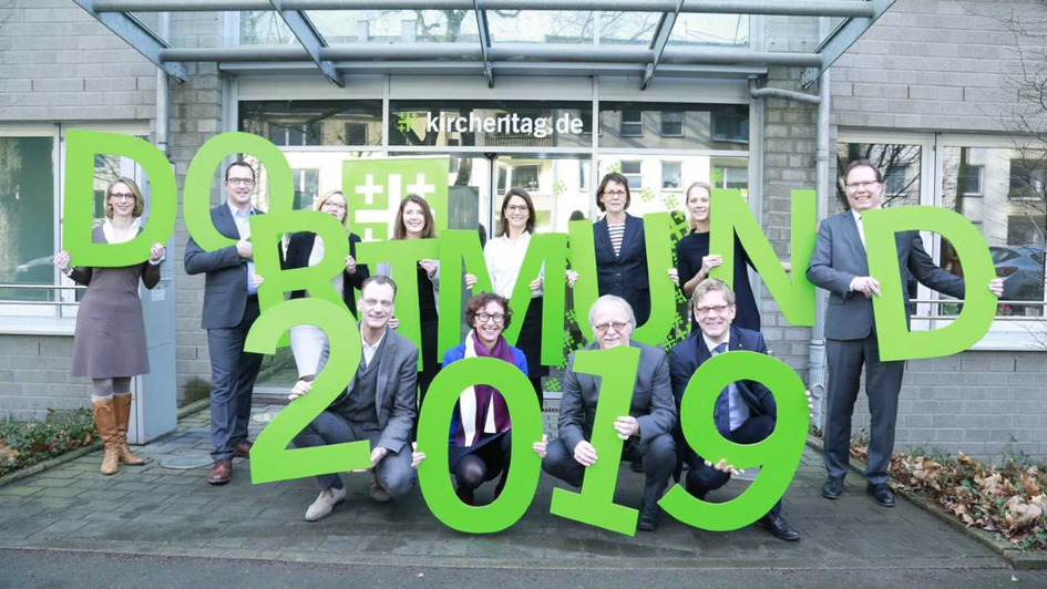 Menschen von der Geschäftsstelle des Deutsche  Evangelischen Kirchentags halten grüne Buchstaben der Worte 'Dortmund 2019'