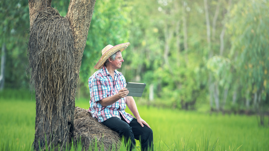 Ein bauer sitzt unter einem Baum am Feld mit einem Tablet in der Hand