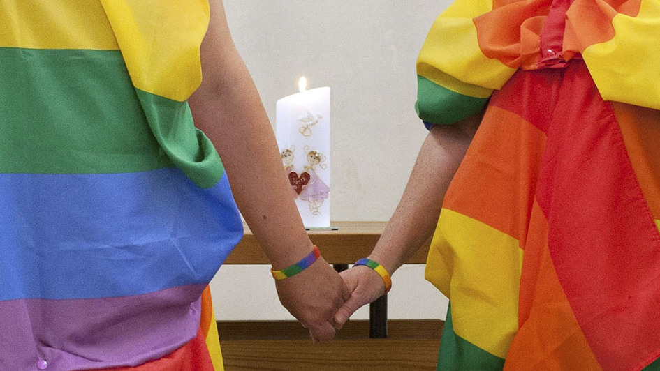 zwei Frauen in Regenbogen-Tüchern halten sich die Hände vor einem Altar