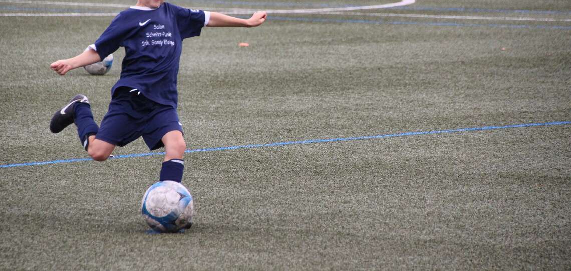 Junge beim Fußballspielen, der gerade im Lauf gegen den Fußball treten will. Der junge trägt ein blaues Trikot und eine blaue Sporthose.
